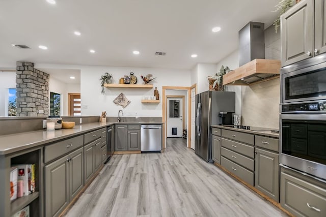 kitchen with gray cabinetry, open shelves, a sink, stainless steel appliances, and wall chimney range hood