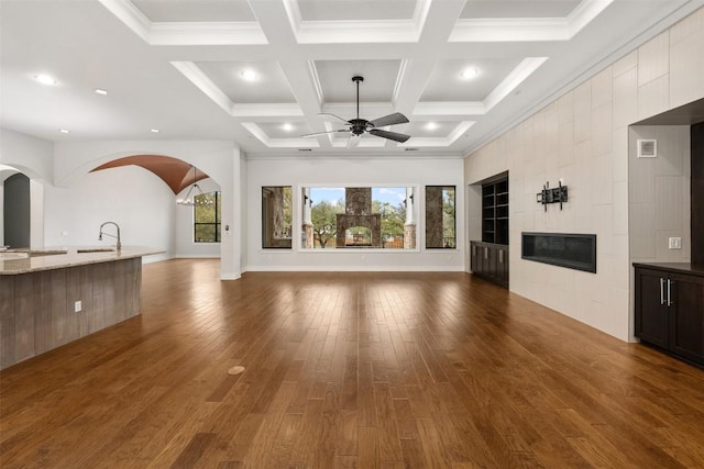 unfurnished living room featuring a ceiling fan, a tiled fireplace, a sink, coffered ceiling, and wood finished floors