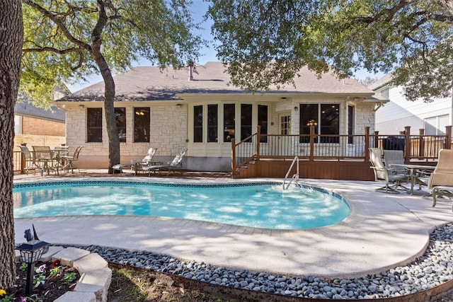 rear view of house with a wooden deck, an outdoor pool, a shingled roof, stone siding, and a patio area