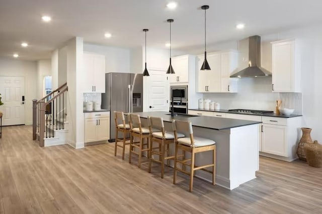 kitchen featuring a breakfast bar, appliances with stainless steel finishes, dark countertops, wall chimney range hood, and light wood-type flooring