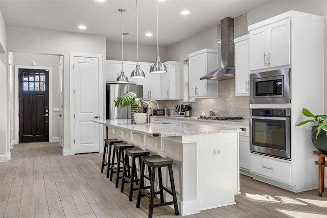 kitchen featuring light wood finished floors, backsplash, wall chimney range hood, appliances with stainless steel finishes, and a sink