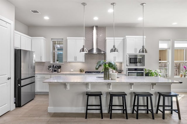 2505 Moreno St, Austin, TX 78723 - kitchen featuring appliances with stainless steel finishes, wall chimney range hood, backsplash, white cabinets, and an island with sink