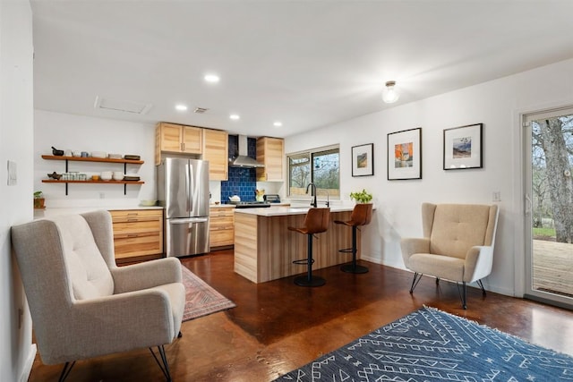 kitchen with light brown cabinets, a peninsula, freestanding refrigerator, wall chimney exhaust hood, and a sink