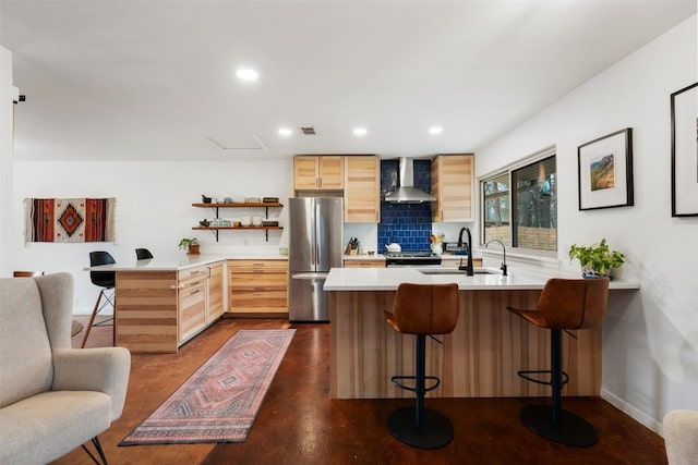 kitchen with a peninsula, a sink, light brown cabinetry, stainless steel appliances, and wall chimney range hood