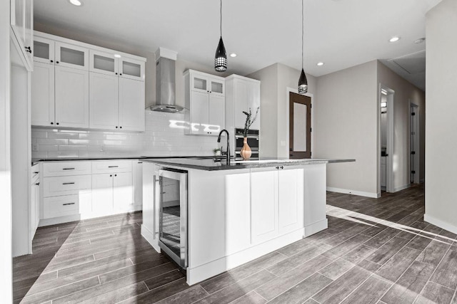 kitchen with beverage cooler, wood finish floors, an island with sink, wall chimney exhaust hood, and backsplash