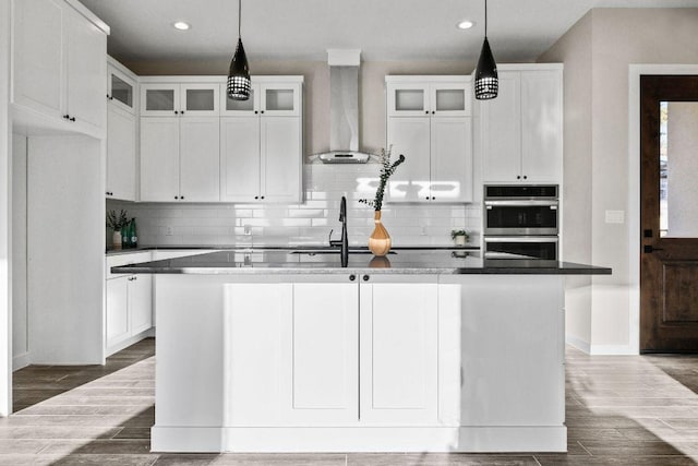 kitchen featuring wall chimney range hood, a center island with sink, white cabinets, and wood tiled floor