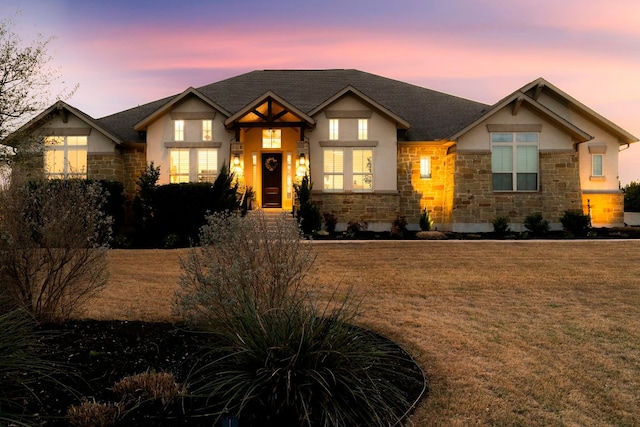 view of front facade with stucco siding, stone siding, a lawn, and a shingled roof