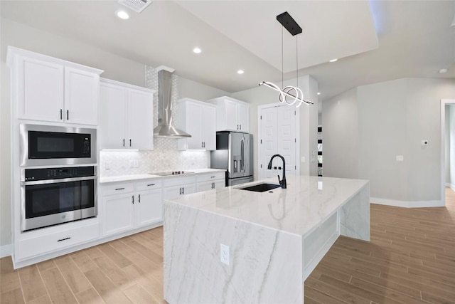 kitchen featuring a kitchen island with sink, a sink, white cabinets, appliances with stainless steel finishes, and wall chimney exhaust hood