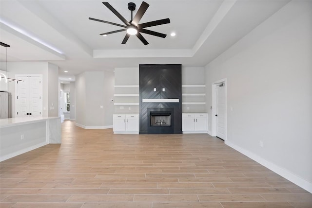 unfurnished living room with light wood finished floors, a large fireplace, baseboards, a tray ceiling, and a ceiling fan