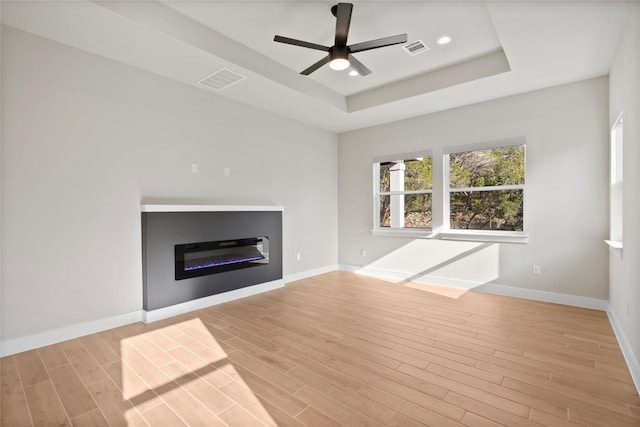 unfurnished living room with baseboards, visible vents, a glass covered fireplace, a raised ceiling, and light wood-type flooring