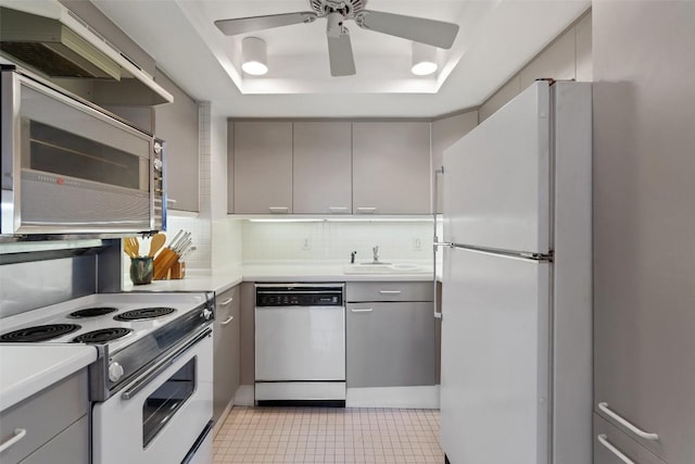 kitchen with gray cabinetry, a tray ceiling, decorative backsplash, white appliances, and a sink