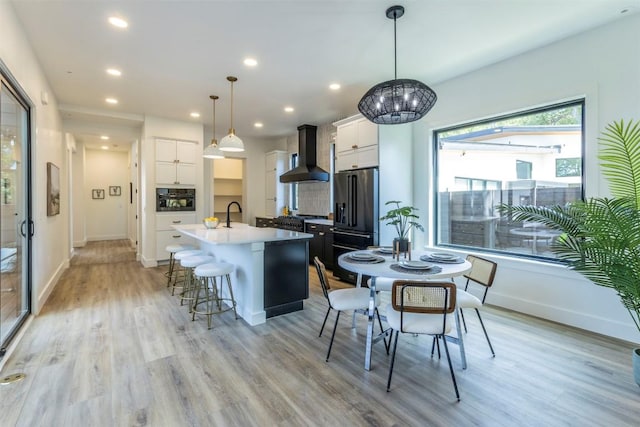 kitchen with light wood-type flooring, black appliances, a center island with sink, a breakfast bar area, and wall chimney range hood