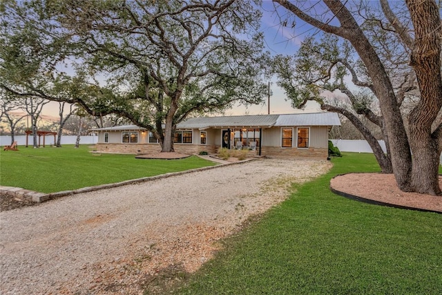 view of front of house featuring a lawn, stone siding, fence, gravel driveway, and metal roof