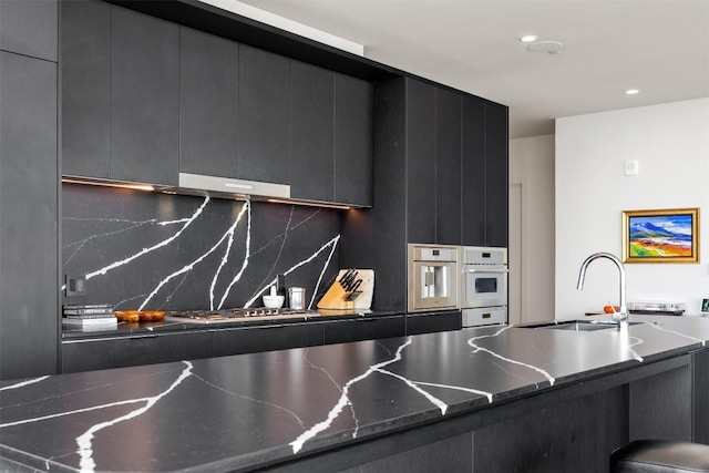 kitchen with modern cabinets, a sink, under cabinet range hood, tasteful backsplash, and dark cabinetry