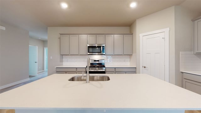 kitchen with gray cabinetry, an island with sink, stainless steel appliances, light countertops, and decorative backsplash