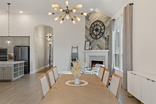 dining space featuring light wood finished floors, high vaulted ceiling, an inviting chandelier, arched walkways, and a lit fireplace