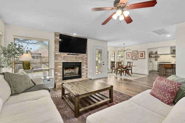 living room with visible vents, recessed lighting, dark wood-style flooring, a brick fireplace, and ceiling fan with notable chandelier