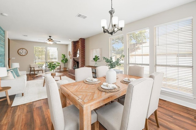 dining room featuring visible vents, ceiling fan with notable chandelier, baseboards, a brick fireplace, and dark wood-style flooring