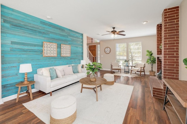 living room featuring dark wood finished floors, stairway, baseboards, ceiling fan, and an accent wall