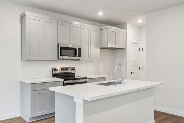 kitchen featuring gray cabinetry, a center island with sink, a sink, light stone counters, and appliances with stainless steel finishes