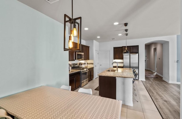 kitchen featuring a sink, light stone counters, arched walkways, dark brown cabinetry, and appliances with stainless steel finishes