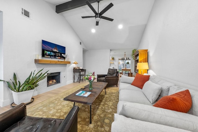 living room featuring a ceiling fan, wood finished floors, visible vents, beam ceiling, and a warm lit fireplace