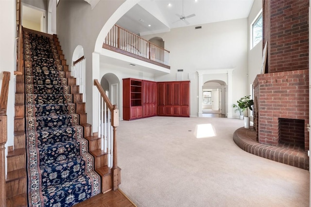 foyer with light carpet, a ceiling fan, stairway, a brick fireplace, and a towering ceiling