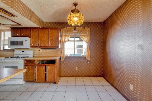 kitchen with wallpapered walls, light tile patterned floors, brown cabinetry, white appliances, and a textured ceiling