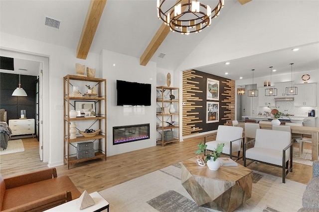 living area with visible vents, an inviting chandelier, light wood-style flooring, a glass covered fireplace, and beamed ceiling