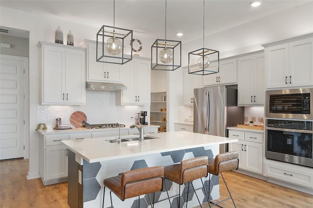 kitchen with light wood finished floors, a kitchen island with sink, under cabinet range hood, appliances with stainless steel finishes, and light countertops