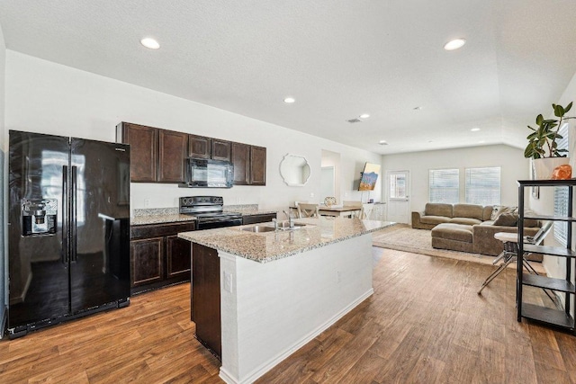 kitchen with a kitchen island with sink, dark brown cabinetry, dark wood-style flooring, black appliances, and a sink