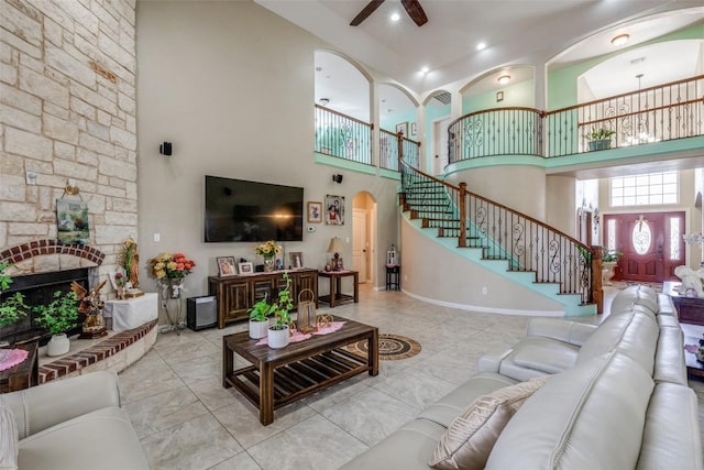 living room featuring a high ceiling, arched walkways, ceiling fan, a stone fireplace, and stairs