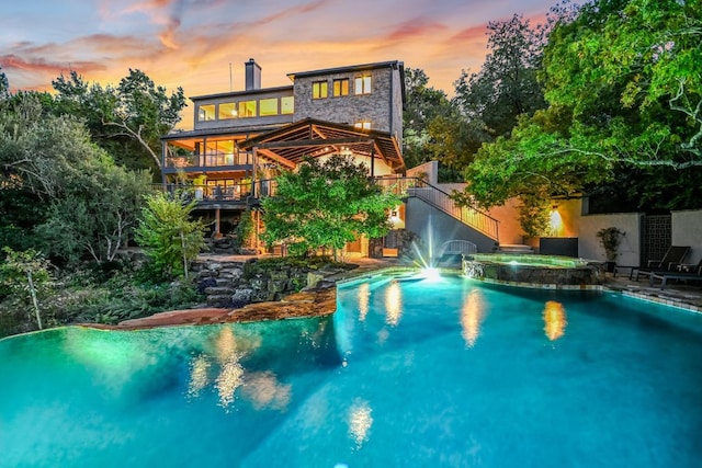 back of house at dusk featuring stone siding, a pool with connected hot tub, a chimney, and stairs