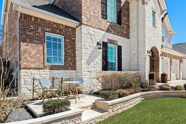 view of side of property with a patio, roof with shingles, an attached garage, stone siding, and brick siding