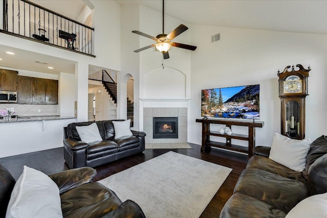 living room with visible vents, dark wood-style flooring, a tiled fireplace, ceiling fan, and stairs