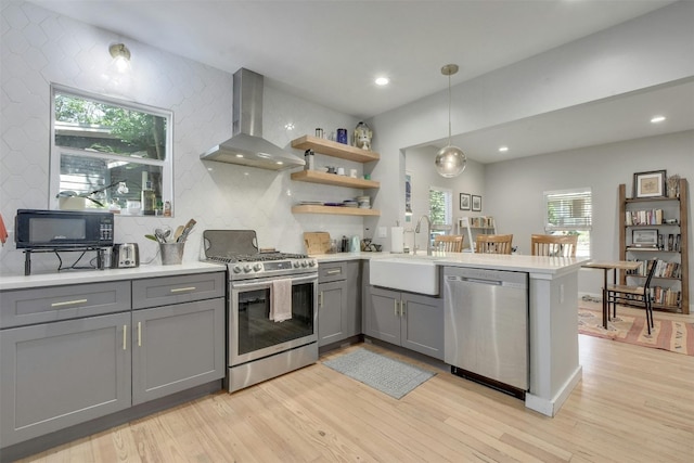 kitchen featuring wall chimney range hood, gray cabinets, appliances with stainless steel finishes, a peninsula, and a sink