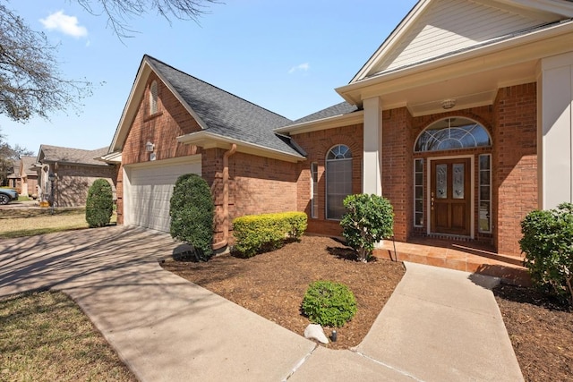 view of exterior entry featuring an attached garage, brick siding, driveway, and a shingled roof