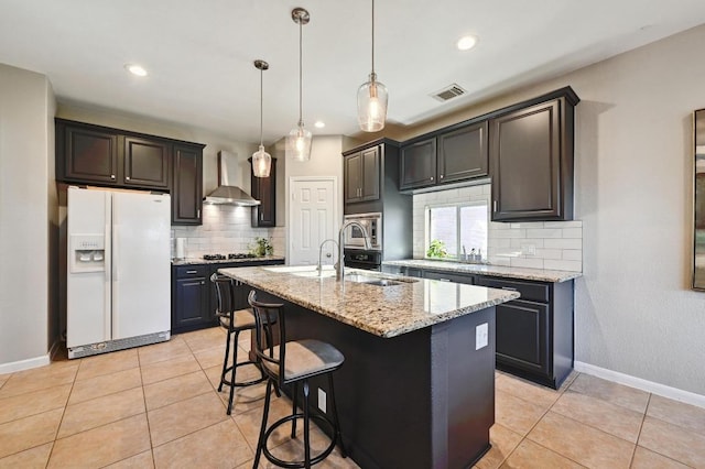 kitchen featuring light stone countertops, visible vents, white refrigerator with ice dispenser, stainless steel microwave, and wall chimney exhaust hood