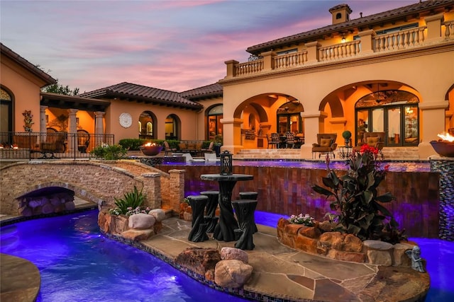 back of house at dusk featuring a tile roof, stucco siding, a balcony, a patio area, and an outdoor pool