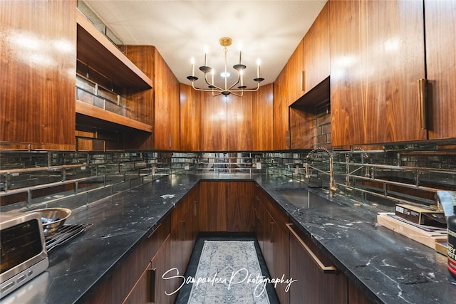 kitchen featuring a sink, open shelves, backsplash, dark stone counters, and an inviting chandelier