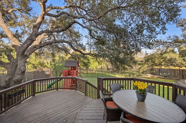 deck with outdoor dining space, a playground, a fenced backyard, and a lawn