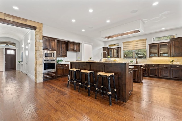 kitchen featuring appliances with stainless steel finishes, an island with sink, dark brown cabinets, a breakfast bar area, and decorative backsplash