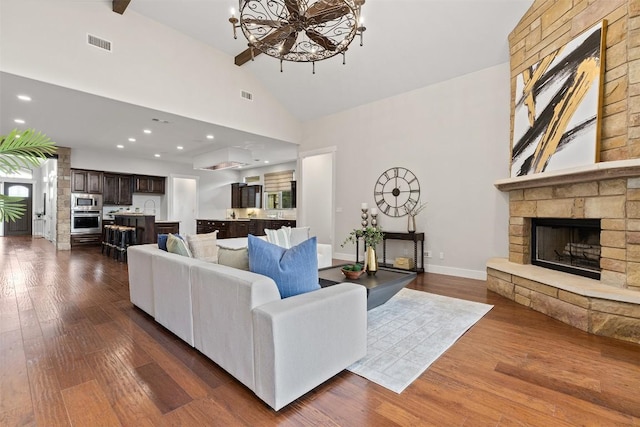 living area featuring a stone fireplace, high vaulted ceiling, dark wood-style flooring, a chandelier, and recessed lighting