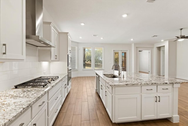 kitchen with a sink, light wood-style floors, stainless steel appliances, wall chimney exhaust hood, and a kitchen island with sink