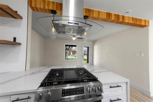 kitchen with light stone countertops, stainless steel gas range, ceiling fan, white cabinetry, and island range hood