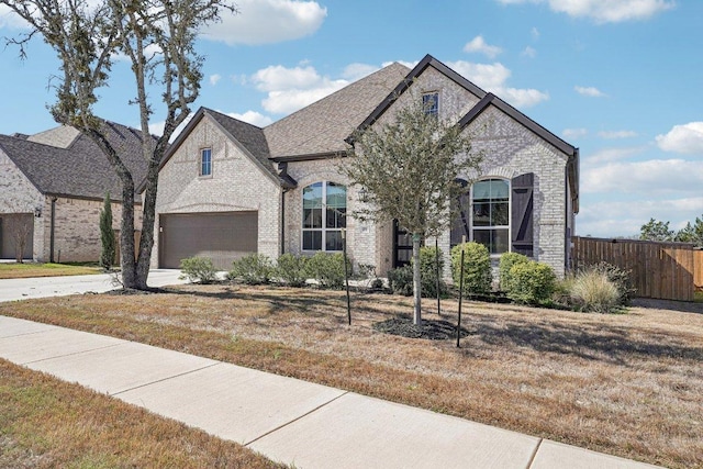 french country inspired facade with fence, driveway, roof with shingles, a garage, and brick siding