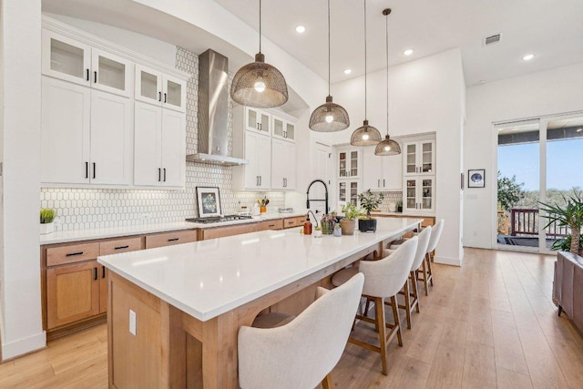 kitchen with light wood-style flooring, gas cooktop, backsplash, wall chimney range hood, and a large island with sink