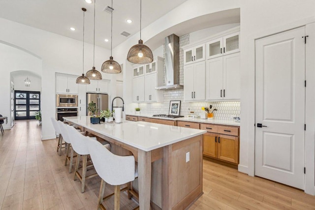 kitchen with arched walkways, stainless steel appliances, decorative backsplash, wall chimney range hood, and light wood-type flooring