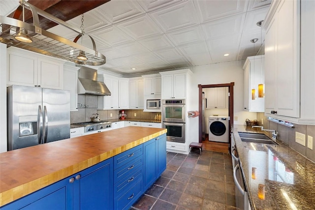 kitchen with blue cabinets, stainless steel appliances, wall chimney range hood, wooden counters, and washer / dryer