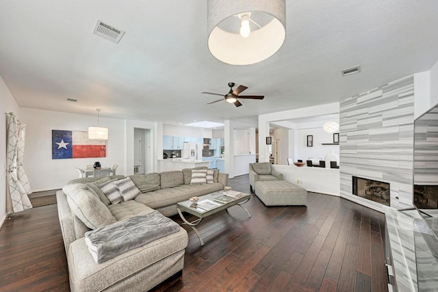 living room with a tiled fireplace, visible vents, dark wood-style flooring, and ceiling fan